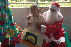 FOTO: Ao lado esquerdo uma Árvore de Natal ao lado direito Papai Noel está entregando um presente a uma aluna, a aluna sorri. 