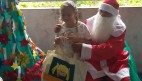 FOTO: Ao lado esquerdo uma Árvore de Natal ao lado direito Papai Noel está entregando um presente a uma aluna, a aluna sorri. 