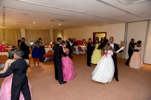 Foto: Ao centro do salão as alunas dançam a valsa com seus pares. Os convidados estão em volta, alguns sentados outros de pé tirando fotos.  