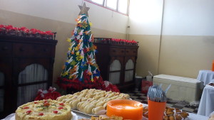 FOTO: A foto mostra os alimentos na mesa principal do buffet.   Ao fundo da foto o foco está para a Árvore de Natal.