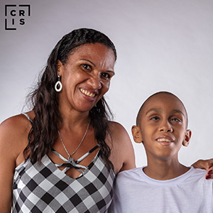 foto na altura do busto, de uma mãe vestida com uma camisa quadriculada, abraçando seu filho, vestido com uma camisa branca. Ambos estão de pé e sorrindo.