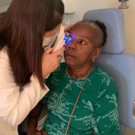 foto de um atendimento oftalmológico, realizado em um dos consultórios do ICB. Nela, uma médica oftalmologista, vestida de jaleco branco, está examinando a visão de uma paciente, que está sentada, vestida com uma camisa verde.