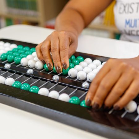 foto de duas mãos, focadas e destacadas, de uma pessoa utilizando o Soroban sobre uma mesa.