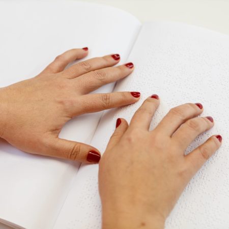 foto de duas mãos, focadas e destacadas, de uma pessoa lendo um livro em Braille sobre uma mesa.