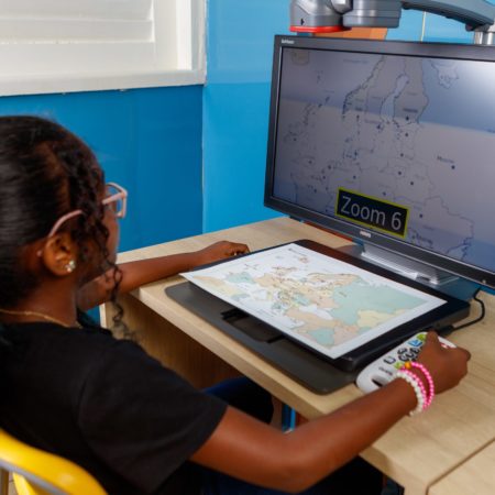 foto de uma criança sentada, em frente a uma tela na Biblioteca do ICB. A menina está utilizando um equipamento que aumenta o zoom, para poder ver uma imagem maior projetada.
