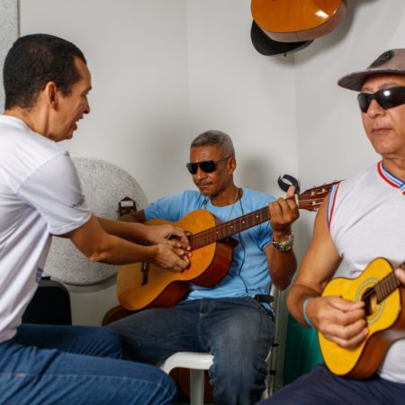 foto dois alunos em uma sala do Centro Musical Imaginasom no ICB. Eles estão sentados, o primeiro do lado esquerdo da foto tocando um violão e outro do lado direito da foto tocando um cavaquinho, sendo orientados por um professor.