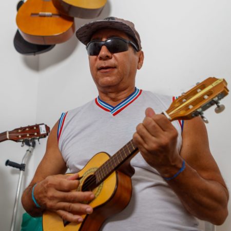 foto um aluno em uma sala do Centro Musical Imaginasom no ICB. Ele está sentado, tocando um cavaquinho, vestido com uma camisa branca e um short azul, com um boné marrom na cabeça e óculos escuro preto. Ao fundo, dois violões e uma guitarra estão pendurados na parede.
