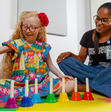 foto de uma criança sentada, ao lado de uma professora de música, em uma das salas do Centro Musical Imaginasom no ICB. A menina está tocando alguns sinos coloridos, em uma aula de iniciação musical.