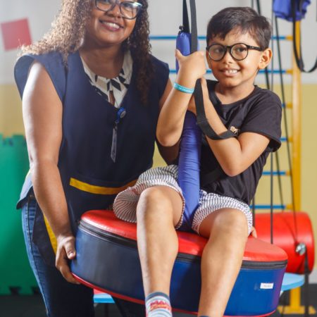 foto de uma criança sentada em um balanço, em uma sala de psicomotricidade do ICB. O menino está vestido com um short claro, uma camisa preta, meias azuis e óculos de cor preta. Ao lado dele, uma professora está de pé, vestida com uma calça jeans, um jaleco azul marinho e utilizando óculos. Ambos estão sorrindo.
