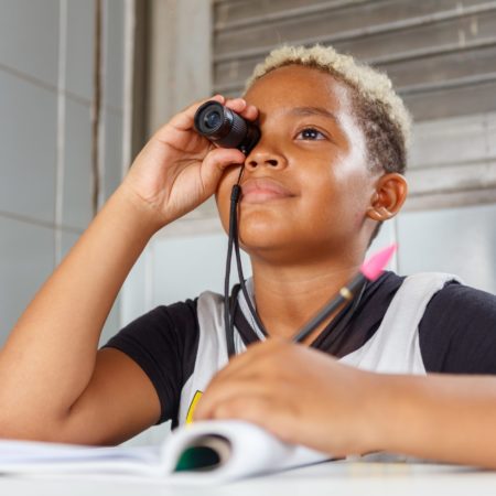 foto de uma criança sentada na altura do busto, em uma sala de aula do ICB. Ele está vestido com uma camisa branca e preta, com um caderno aberto e uma caneta na mão. Ela está segurando um recurso óptico no olho direito.