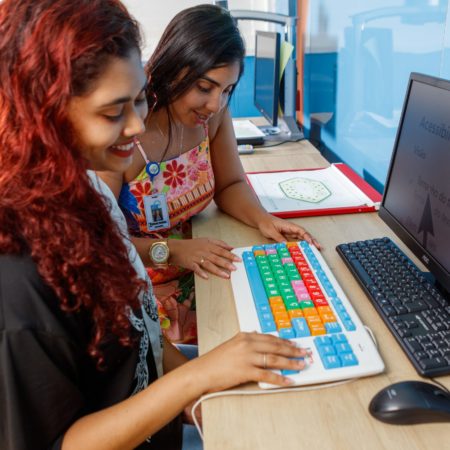 foto de uma mulher sentada, ao lado de uma professora, em frente a um computador na Biblioteca do ICB. Elas estão olhando para um teclado colorido, utilizado para dar contraste de cores, e na tela está uma imagem com um aumento de fontes, para uma melhor visualização e acessibilidade.