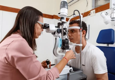 foto de um atendimento oftalmológico, realizado em um dos consultórios do ICB. Nela, uma médica oftalmologista, vestida de jaleco rosa, está examinando a visão de um paciente, que está sentado em frente a um aparelho, vestido com uma camisa branca.