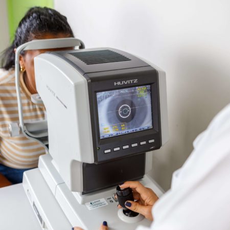 foto de um atendimento oftalmológico, realizado em um dos consultórios do ICB. Nela, uma médica oftalmologista, vestida de jaleco branco, está realizando um exame computadorizado em uma paciente, que está sentada, vestida com uma camisa listrada.