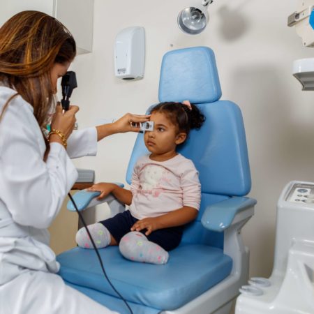 foto de um atendimento oftalmológico, realizado em um dos consultórios do ICB. Nela, uma médica oftalmologista, vestida de jaleco branco, está examinando a visão de uma criança, que está sentada, vestida com uma camisa rosa e short preto.