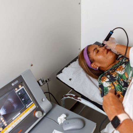 foto de um atendimento oftalmológico, realizado em um dos consultórios do ICB. Nela, uma médica oftalmologista, vestida de jaleco branco, está realizando um exame computadorizado em uma paciente, que está deitada em uma maca, de olhos fechados, vestida com uma camisa floral.