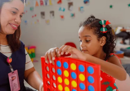 foto em uma das salas do ICB, onde a Terapeuta Ocupacional, vestida com uma camisa branca e um jaleco azul marinho, está sentada ao lado de uma menina, que está realizando uma atividade, manuseando um brinquedo de cor vermelha, azul e amarela. Ela está com um vestido vermelho com alguns detalhes de cor verde e branco e um laço no cabelo.