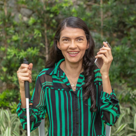 foto de uma mulher, na altura do busto, segurando uma bengala com a mão direita e com a mão esquerda um celular. Ela está vestida com uma camisa verde, sorrindo. Ao fundo um jardim com plantas.
