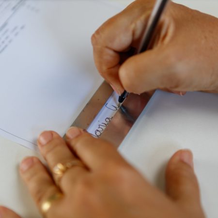 foto de duas mãos, focadas e destacadas, de uma pessoa assinando o nome com a ajuda de um “assinador”.