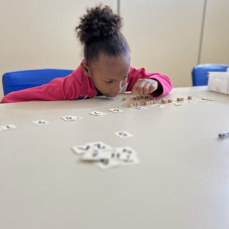 foto de uma criança sentada na altura do busto, em uma sala de aula do ICB. Ela está vestida com um casaco rosa, realizando uma atividade sobre uma mesa, com papéis e pequenos blocos de madeira.