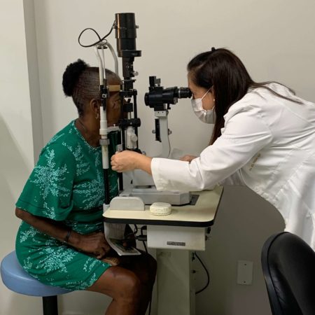 foto de um atendimento oftalmológico, realizado em um dos consultórios do ICB. Nela, uma médica oftalmologista, vestida de jaleco branco, está examinando a visão de um paciente, que está sentado em frente a um aparelho, utilizando um vestido verde.