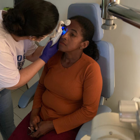 foto de um atendimento oftalmológico, realizado em um dos consultórios do ICB. Nela, uma médica oftalmologista, vestida com uma camisa branca, está examinando a visão de uma paciente, que está sentada, vestida com uma camisa marrom e uma calça vermelha.