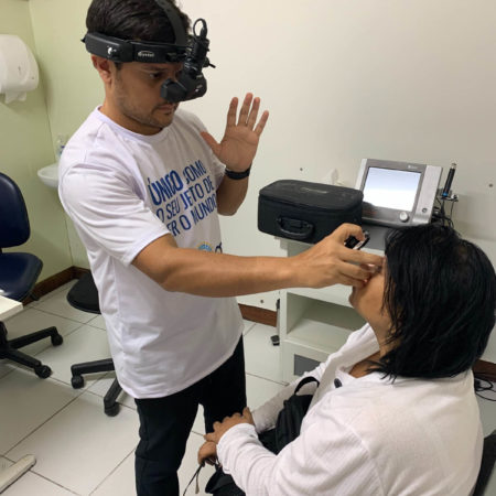 foto de um atendimento oftalmológico, realizado em um dos consultórios do ICB. Nela, um médico oftalmologista, vestido com uma camisa branca e calça jeans, está examinando a visão de uma paciente, que está sentada, vestindo uma camisa branca e calça jeans.