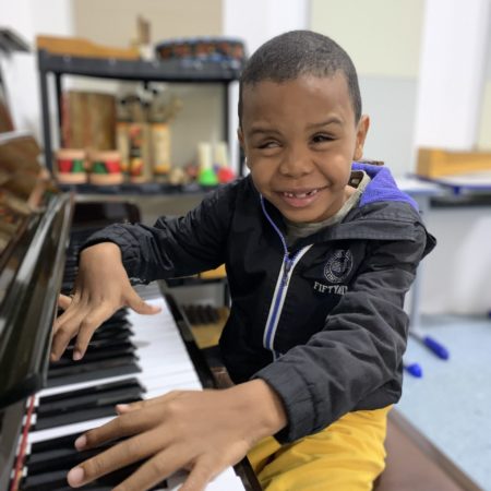 foto de um menino, sentado em uma sala de iniciação musical do ICB. Ele está sentado e sorrindo, tocando um piano, vestido com um short amarelo e um casaco preto.