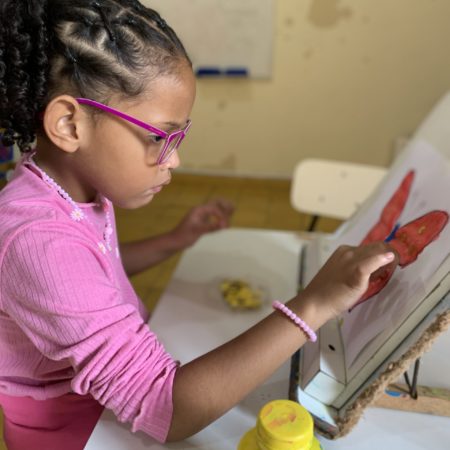 foto de uma criança sentada na altura da cintura, em uma sala de aula do ICB. Ela está vestida com uma camisa rosa, óculos rosa e cabelos trançados. Ela está realizando uma atividade sobre a mesa, colorindo um desenho com tinta.